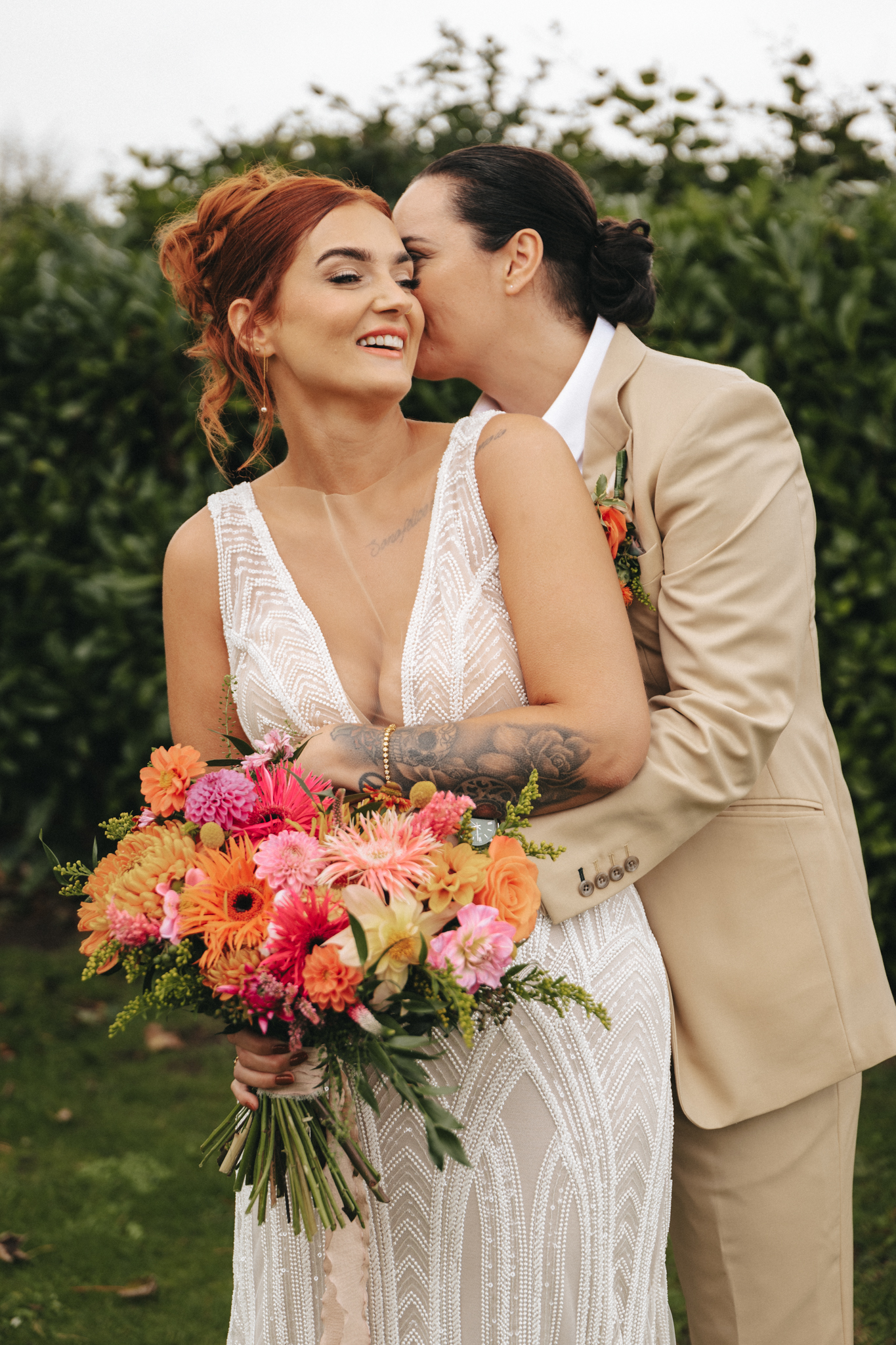 A couple embraces outdoors on their wedding day. One person, with red hair and a tattoo on their forearm, is wearing a white sleeveless wedding gown and holding a vibrant bouquet of flowers. The other, with long dark hair tied back, is dressed in a beige suit and is leaning in for a kiss on the cheek. They stand in front of lush greenery, capturing the magic of their wedding © Aimee Lince Photography