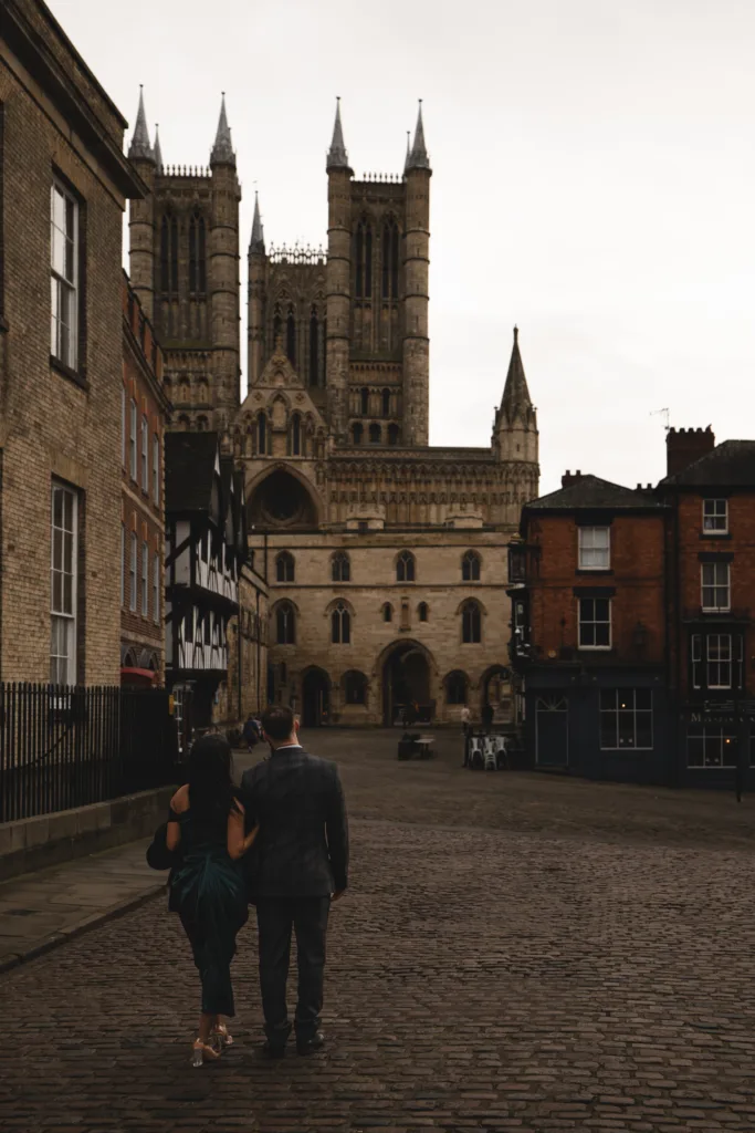 A couple enjoying their engagement session stroll on a picturesque cobbled street in Lincoln, with a majestic cathedral adorning the background.