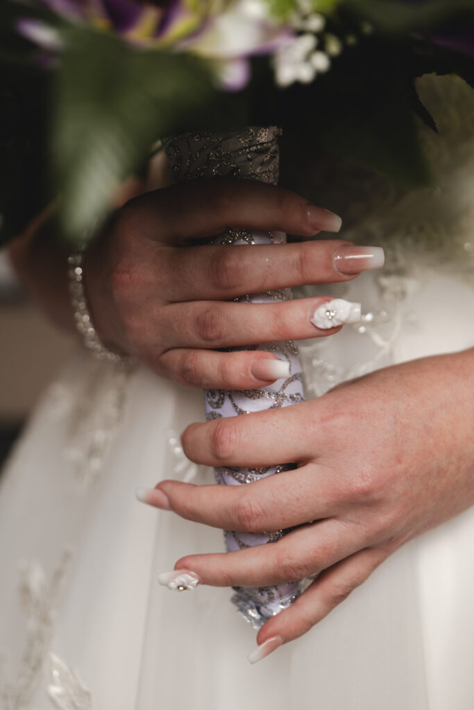 A close-up shows a person in a white lace wedding dress holding a bejeweled bouquet handle with both hands. The person has manicured nails with a white and pink French tip design adorned with small white flowers and pearls. At The Humber Royal Hotel, the bouquet's green leaves and white flowers peek through elegantly. © Aimee Lince Photography