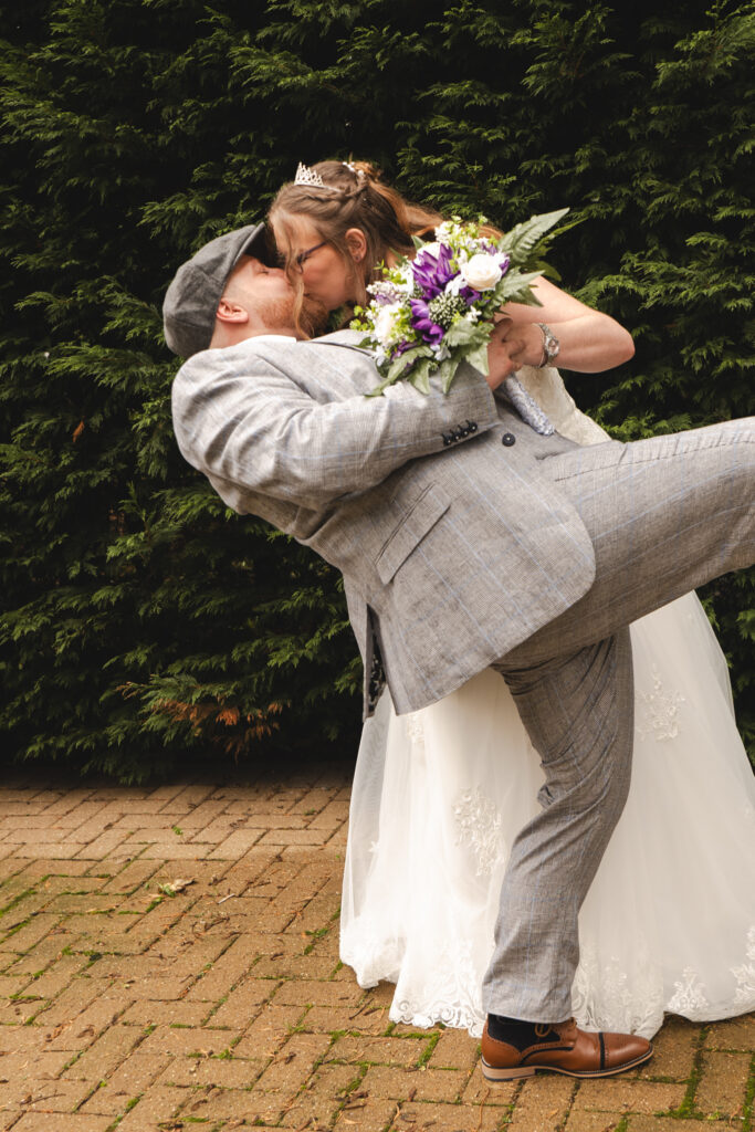 A newlywed couple shares a kiss outdoors at The Humber Royal Hotel. The groom, dressed in a gray suit and cap, leans back while holding the bride, who wears a white gown and tiara. She holds a bouquet of purple and white flowers. Both stand on a brick pathway, with a dense green hedge as the backdrop. © Aimee Lince Photography