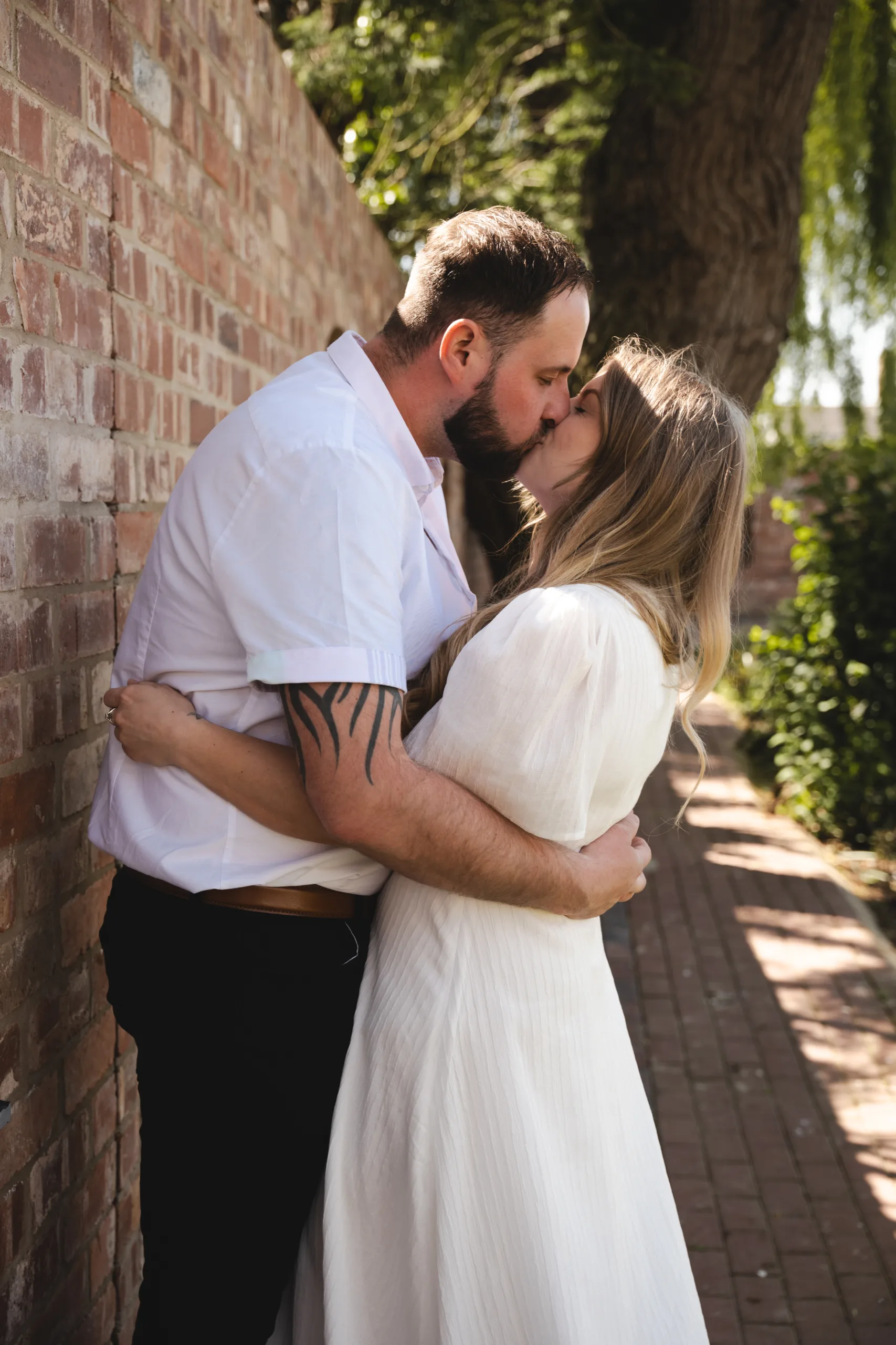 A couple in engagement photography attire share a romantic kiss in front of a brick wall.