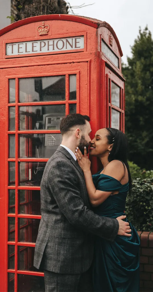 A couple embraces in front of a vintage red telephone booth, capturing the perfect engagement photography moment. The man, in a gray suit, and the woman, in a dark green dress, share a tender smile as she gently holds his face. Greenery and part of Lincoln's charming architecture are visible behind them. © Aimee Lince Photography - Wedding photographer in Lincolnshire, Yorkshire & Nottinghamshire