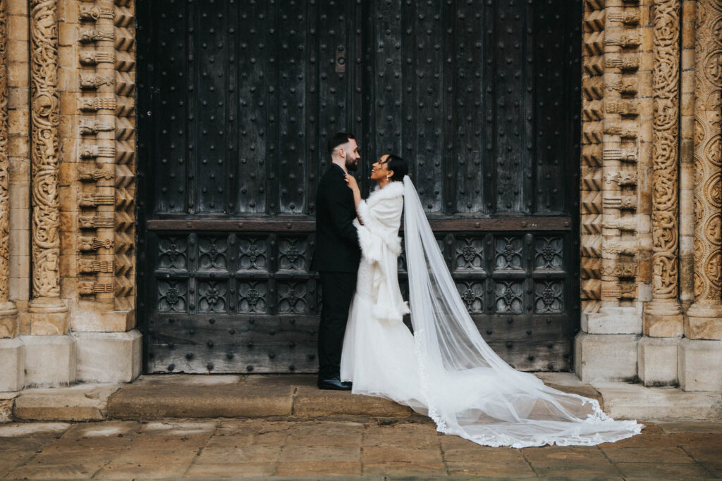Lincoln Cathedral Wedding Photoshoot - Ed & Makda © Aimee Lince Photography 02-12-23 2222 of 4040 A bride and groom stand closely in front of a large, ornate wooden door at Lincoln Cathedral. The bride wears a long white dress with a sheer, flowing veil and holds the groom's lapel. The groom wears a black suit. The door is flanked by intricately carved stone columns. © Aimee Lince Photography