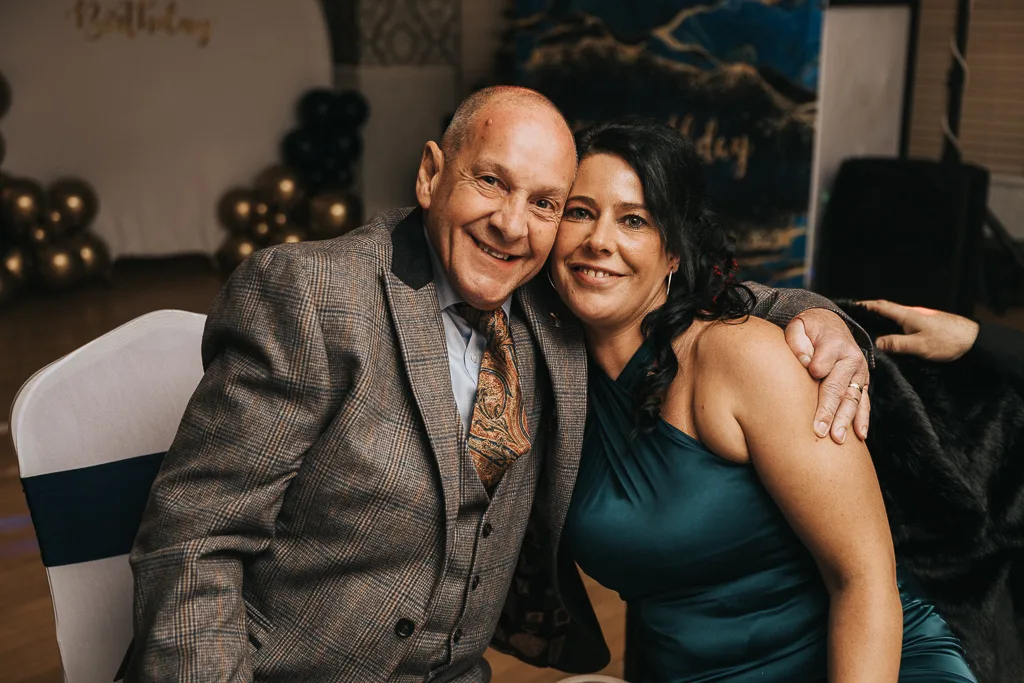 An older man in a plaid suit and patterned tie smiles while seated next to a woman in a teal dress with long dark hair. Arm in arm, they enjoy the masquerade-themed party, surrounded by black and gold balloons. The atmosphere is warm and celebratory, perfect for capturing memorable photography. © Aimee Lince Photography - Wedding photographer in Lincolnshire, Yorkshire & Nottinghamshire
