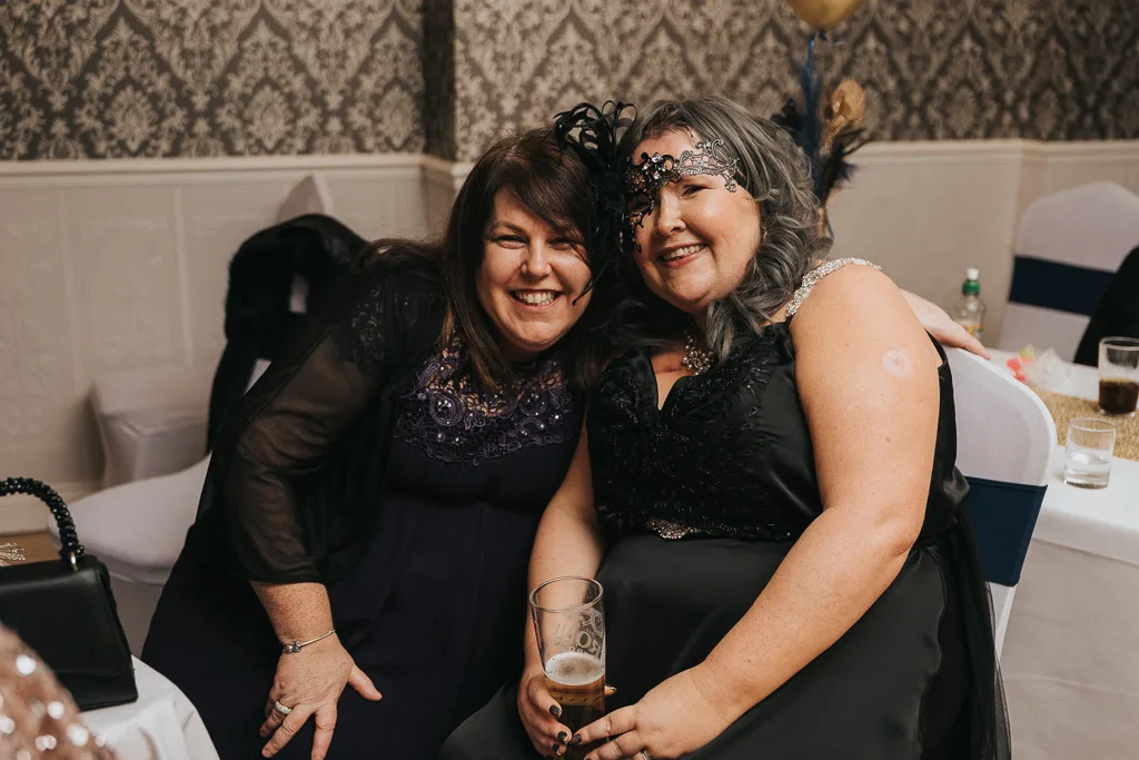Two women in black dresses are sitting and smiling at the camera during a lively party. One wears a feathered headpiece, exuding a masquerade vibe, and holds a drink. They're indoors, with patterned wallpaper and an elegant setting featuring a wrapped bottle and glasses on the table nearby. © Aimee Lince Photography - Wedding photographer in Lincolnshire, Yorkshire & Nottinghamshire