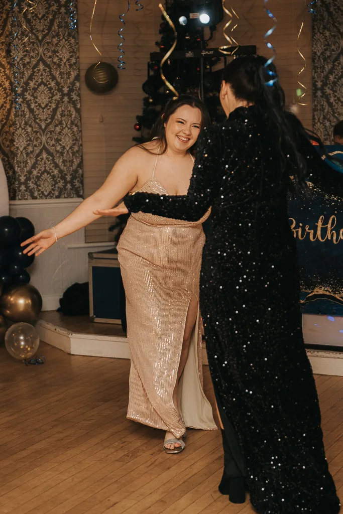 Two women are joyfully hugging on a dance floor at a surprise party. The woman on the left wears a shimmering beige dress, while the woman on the right dazzles in a black sequined dress. The background, adorned with balloons, streamers, and a "Birthday" sign, enhances the festive atmosphere. © Aimee Lince Photography - Wedding photographer in Lincolnshire, Yorkshire & Nottinghamshire