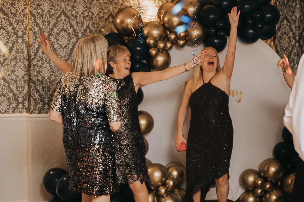 Three women in sparkly dresses dance joyfully near an arch of black and gold balloons, embodying the spirit of a masquerade. The backdrop features elegant, patterned wallpaper. One woman has her arm raised, smiling widely, capturing a moment of surprise and joy in perfect photography. © Aimee Lince Photography - Wedding photographer in Lincolnshire, Yorkshire & Nottinghamshire