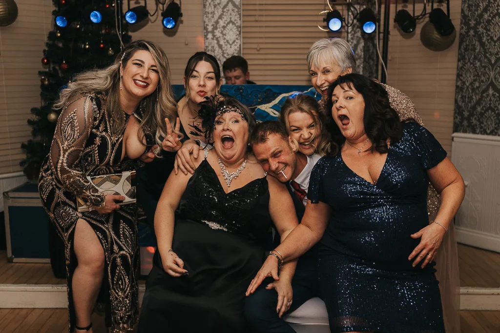 A group of six people pose exuberantly indoors, capturing the essence of party joy. Three women kneel behind a man and two seated women, all smiling broadly with some flashing peace signs. Sparkly dresses and a suit add to their festive spirit, as a Christmas tree and lights frame this joyful moment in photography. © Aimee Lince Photography - Wedding photographer in Lincolnshire, Yorkshire & Nottinghamshire