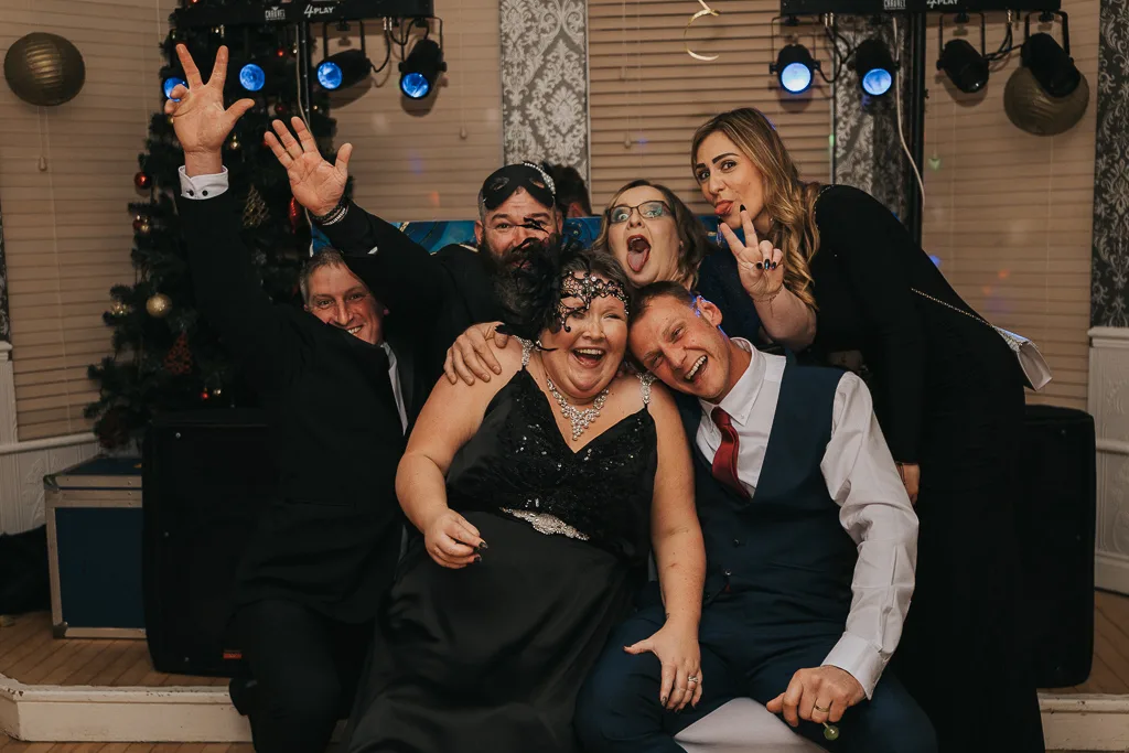 A group of six people poses joyfully at a birthday party. A woman in a black dress and a man in a blue suit are seated in front, leaning together. Four others stand behind them, smiling and making playful gestures. Photography captures the festive scene, with a Christmas tree and sound equipment visible in the background. © Aimee Lince Photography - Wedding photographer in Lincolnshire, Yorkshire & Nottinghamshire