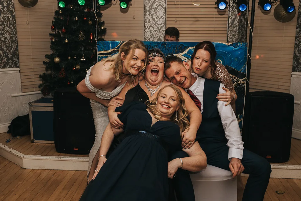 A group of six people are joyfully posing indoors, exuding a spirited party vibe. Two women are seated in front, with three women and one man standing behind them. Everyone is smiling and clad in semi-formal attire. A decorated Christmas tree and large speakers complete the festive scene. © Aimee Lince Photography - Wedding photographer in Lincolnshire, Yorkshire & Nottinghamshire