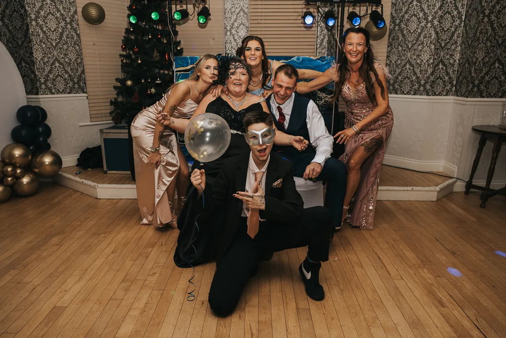A group of six people in formal attire pose joyfully at a masquerade-themed party. A young man kneels in the front, holding a balloon and wearing a mask. Behind him, five adults sit and stand closely, smiling. A Christmas tree with decorations and colorful lights is visible in the background, capturing festive photography moments. © Aimee Lince Photography - Wedding photographer in Lincolnshire, Yorkshire & Nottinghamshire