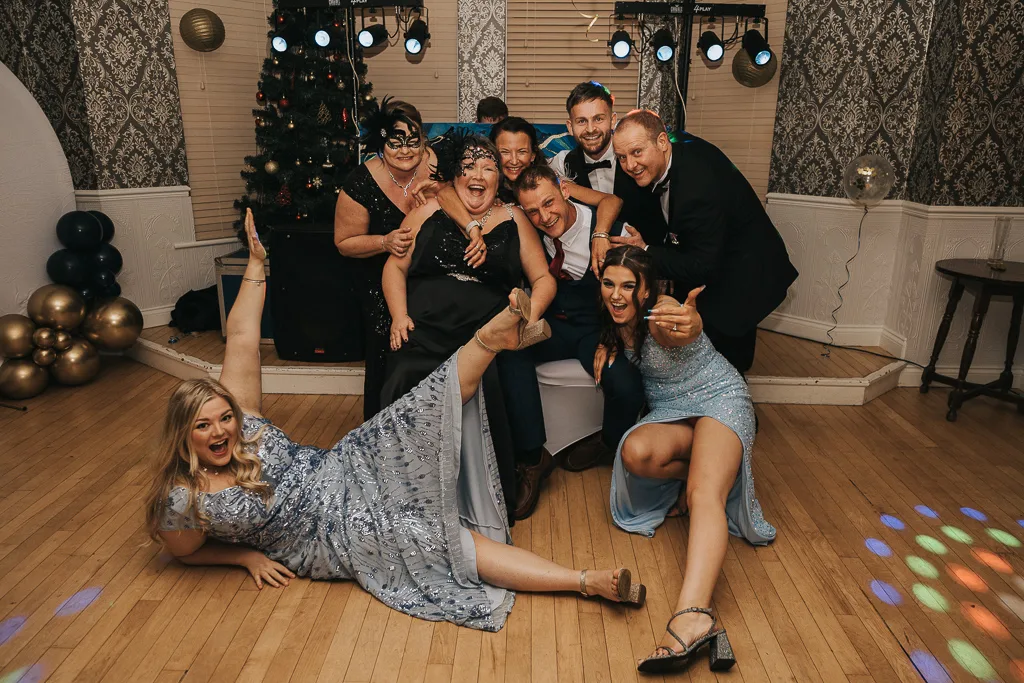 A group of eight people in playful poses at a party surprises everyone with their lively energy. Dressed in formal attire, with the two women in front lying on the floor in light blue dresses, they gather near a Christmas tree under decorative lights, creating perfect photography moments on the festive scene. © Aimee Lince Photography - Wedding photographer in Lincolnshire, Yorkshire & Nottinghamshire