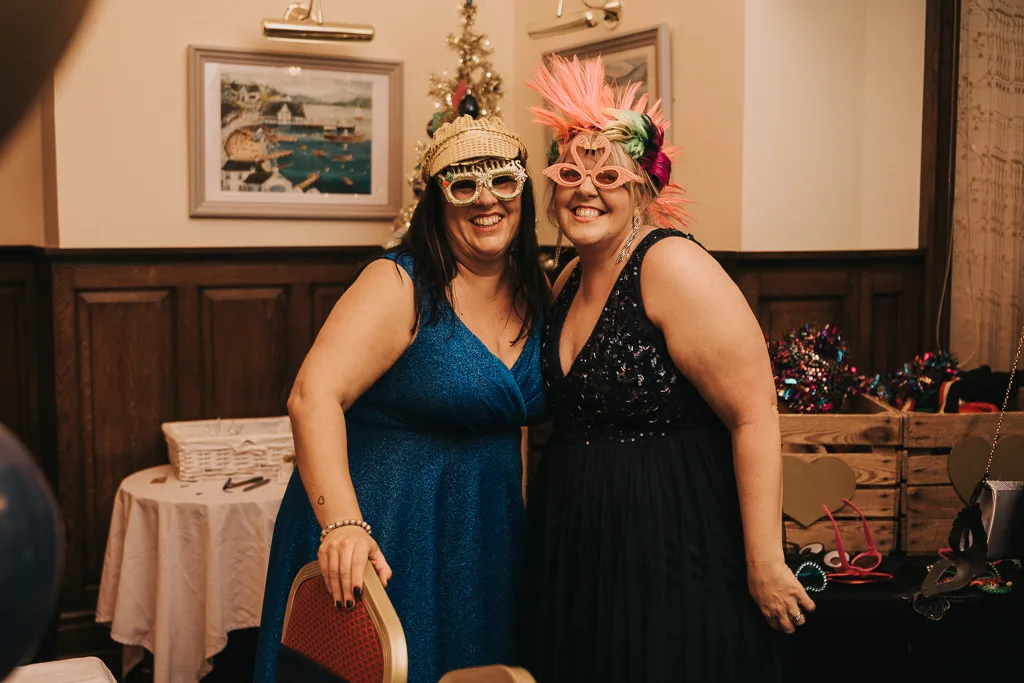Two women in blue dresses pose at a surprise party. They wear playful accessories: oversized glasses, one with feathers and the other with a straw hat. Behind them are a wooden wall, framed pictures, a small decorated Christmas tree, and tables with scattered party items. © Aimee Lince Photography - Wedding photographer in Lincolnshire, Yorkshire & Nottinghamshire