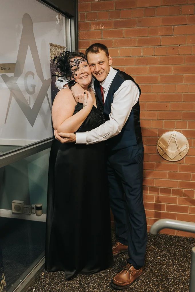 A smiling couple embraces in front of a brick wall, as if ready for a masquerade. The woman wears a black dress with a decorative fascinator; the man sports a dark suit and red tie. Behind them, a large window displays the word “Gents” and hints at a secret birthday surprise. © Aimee Lince Photography - Wedding photographer in Lincolnshire, Yorkshire & Nottinghamshire