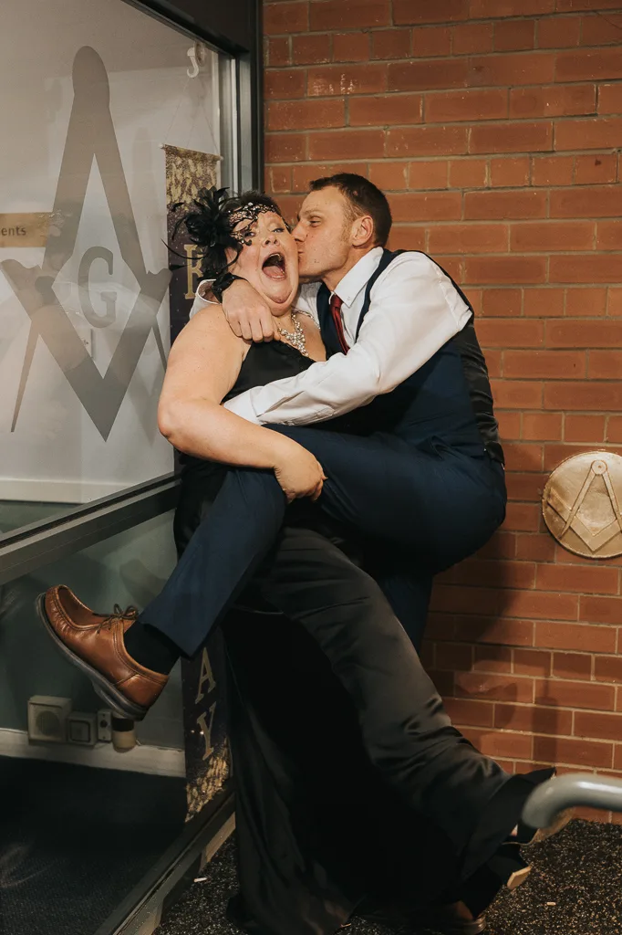 A man in a blue suit and red tie joyfully kisses a laughing woman in a black dress, flower headpiece, and feathered mask at the masquerade party. She lifts her leg playfully as they pose by a brick wall adorned with a Masonic symbol and blue banner. The mood captured in the photography is festive and lively. © Aimee Lince Photography - Wedding photographer in Lincolnshire, Yorkshire & Nottinghamshire