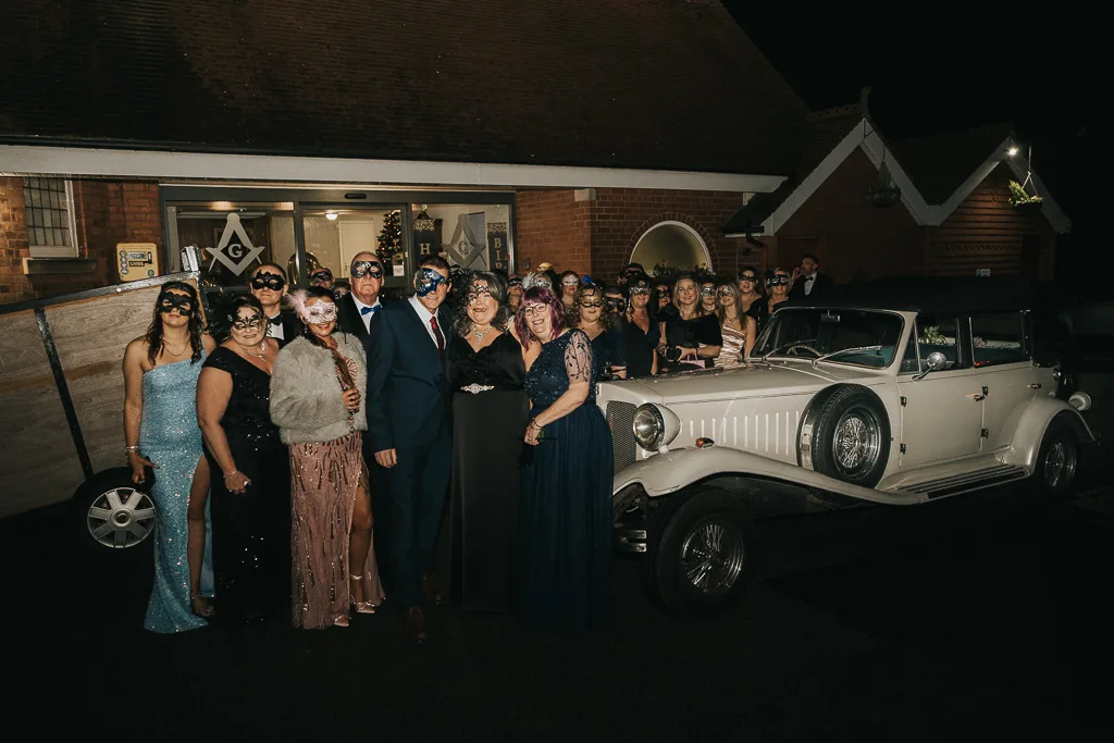 A large group of people, dressed in formal evening wear and masquerade masks, pose beside a vintage white car at night. The setting appears festive, with a building in the background. This enchanting photography captures the group's suits and elegant gowns perfectly, creating a glamorous atmosphere. © Aimee Lince Photography - Wedding photographer in Lincolnshire, Yorkshire & Nottinghamshire