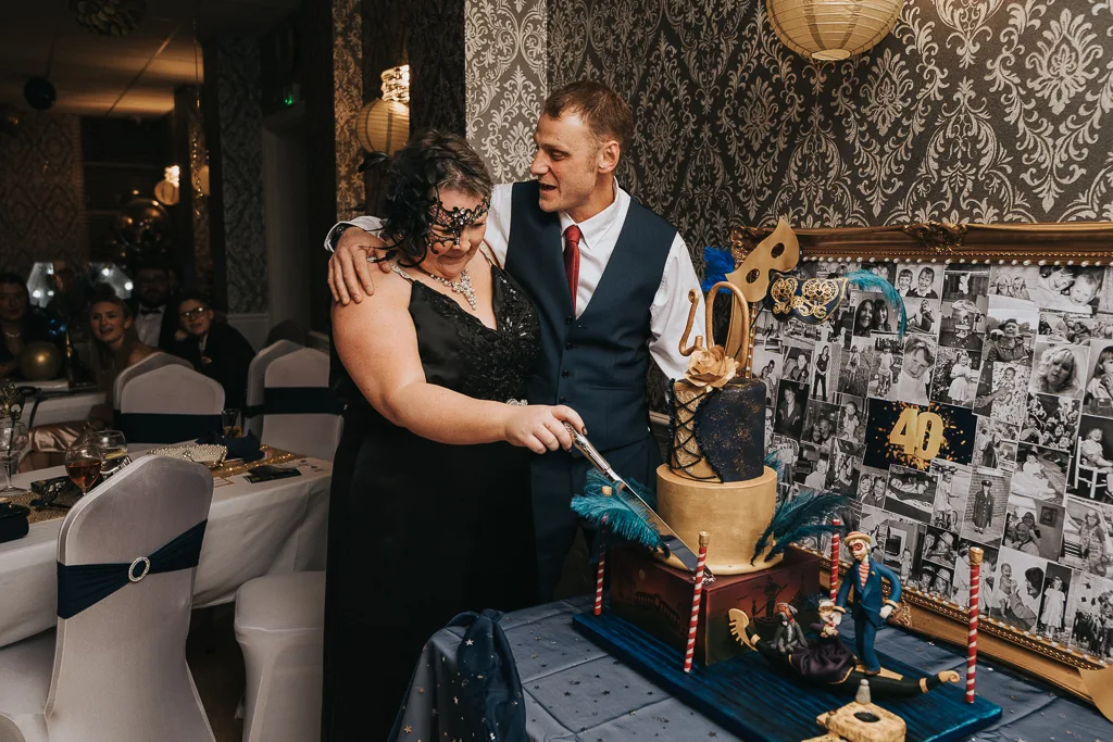 A couple stands by a blue and gold themed cake at the masquerade birthday party. The woman, in a black dress and mask, cuts the cake with the suited man beside her. Photography captures the glittery "40" among collage wall photos as guests sit at tables in a dimly lit room. © Aimee Lince Photography - Wedding photographer in Lincolnshire, Yorkshire & Nottinghamshire