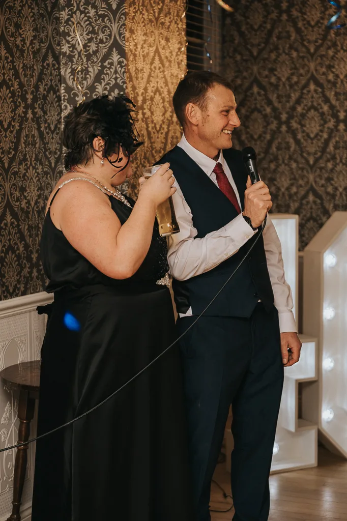 A person in a black dress and feathered mask holds a drink while standing next to someone in a suit with a red tie, speaking into a microphone. They are in a room with ornate wallpaper and illuminated letters, evoking the festive atmosphere of a surprise masquerade party. © Aimee Lince Photography - Wedding photographer in Lincolnshire, Yorkshire & Nottinghamshire