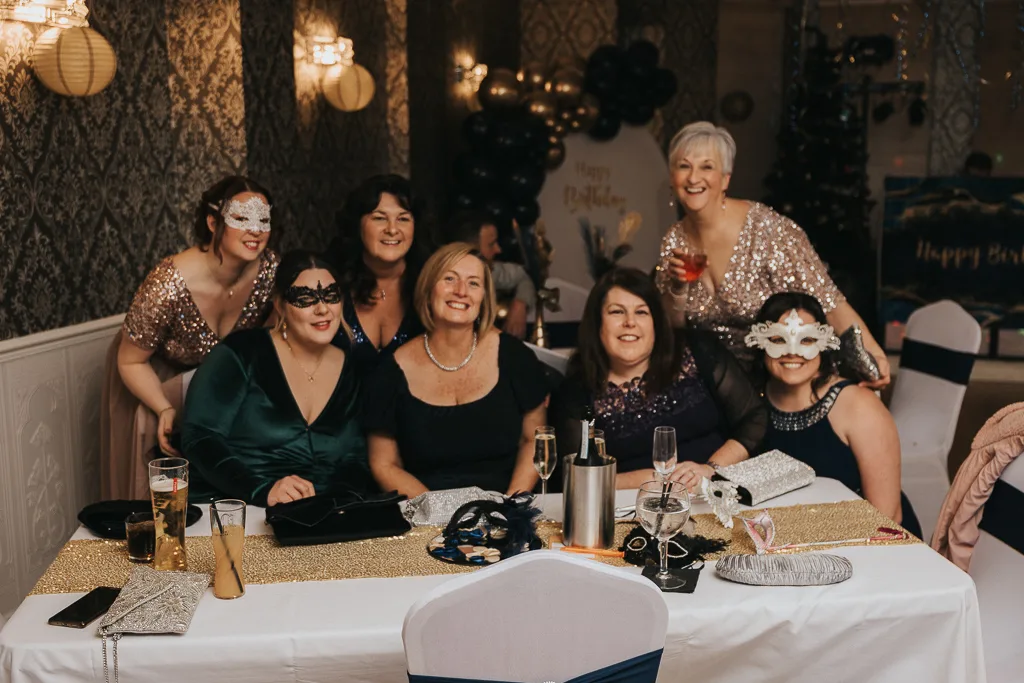 Seven women dressed in formal wear and masks smile at a masquerade party. Four are seated at a table with drinks and purses, while three stand behind them. The room is decorated with patterned wallpaper, balloon arrangements, and dim lighting, creating a festive atmosphere. © Aimee Lince Photography - Wedding photographer in Lincolnshire, Yorkshire & Nottinghamshire