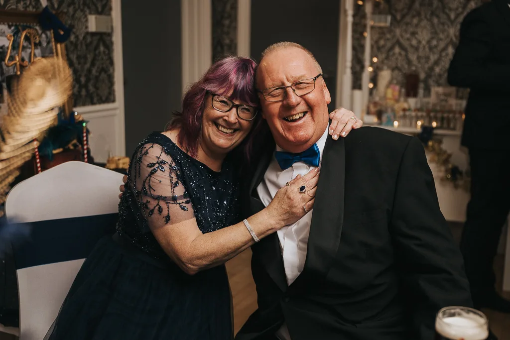 An older couple joyfully poses together indoors, captured in lively photography. The woman in a navy blue, sequin-adorned masquerade dress laughs, adjusting the man's tuxedo lapel. He beams in a black tux and blue bowtie. Festive party decor sets the backdrop for their happiness. © Aimee Lince Photography - Wedding photographer in Lincolnshire, Yorkshire & Nottinghamshire
