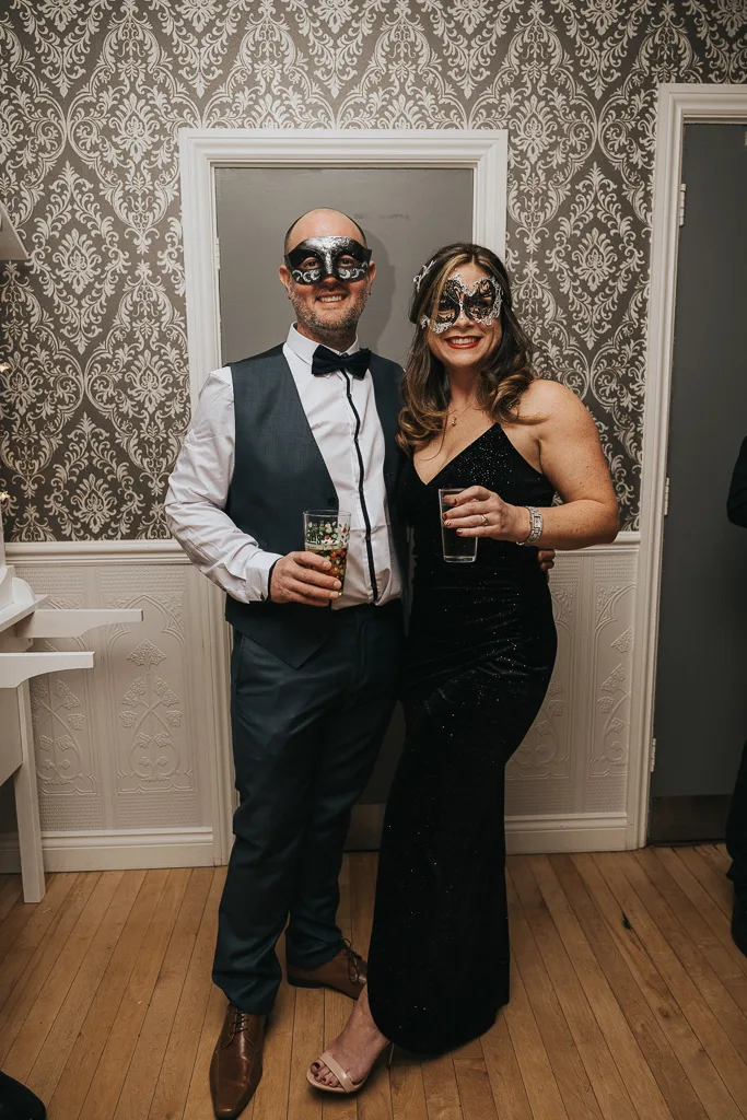 A man and woman in masquerade masks pose stylishly at a lively party. The man sports a gray suit with a bow tie, drink in hand, while the woman dazzles in a black dress. They stand on a wooden floor against elegant patterned wallpaper, capturing the essence of sophisticated event photography. © Aimee Lince Photography - Wedding photographer in Lincolnshire, Yorkshire & Nottinghamshire