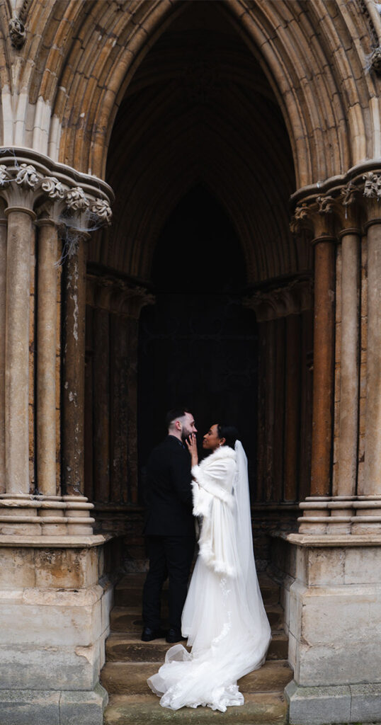 A couple stands in a romantic embrace under the gothic archway of Lincoln Cathedral. The bride, in a long white veil and gown with a fur wrap, gazes up at the groom, who wears a black suit. The stone pillars and intricate design create a dramatic, historic background. © Aimee Lince Photography