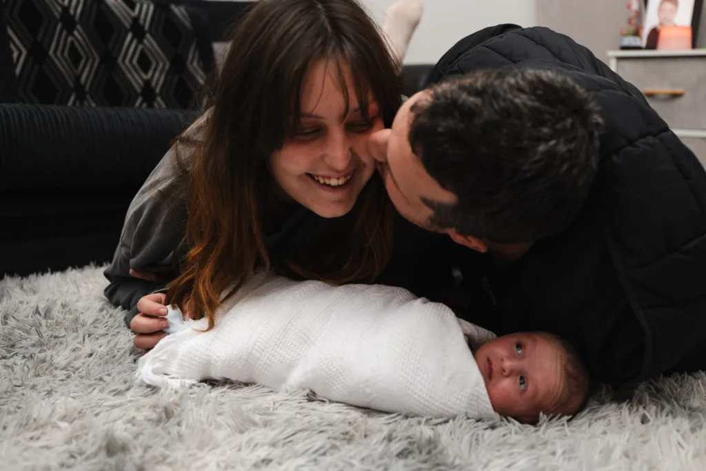 A man and woman tenderly kissing their newborn baby in a cozy living room.