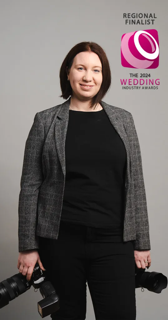 A person with shoulder-length brown hair smiles, holding two cameras. They're wearing a gray plaid blazer over a black shirt and pants. A "Regional Finalist" badge from the 2024 Wedding Industry Awards adorns the top right corner, showcasing their achievement and feedback excellence against a plain gray background. © Aimee Lince Photography - Wedding photographer in Lincolnshire, Yorkshire & Nottinghamshire