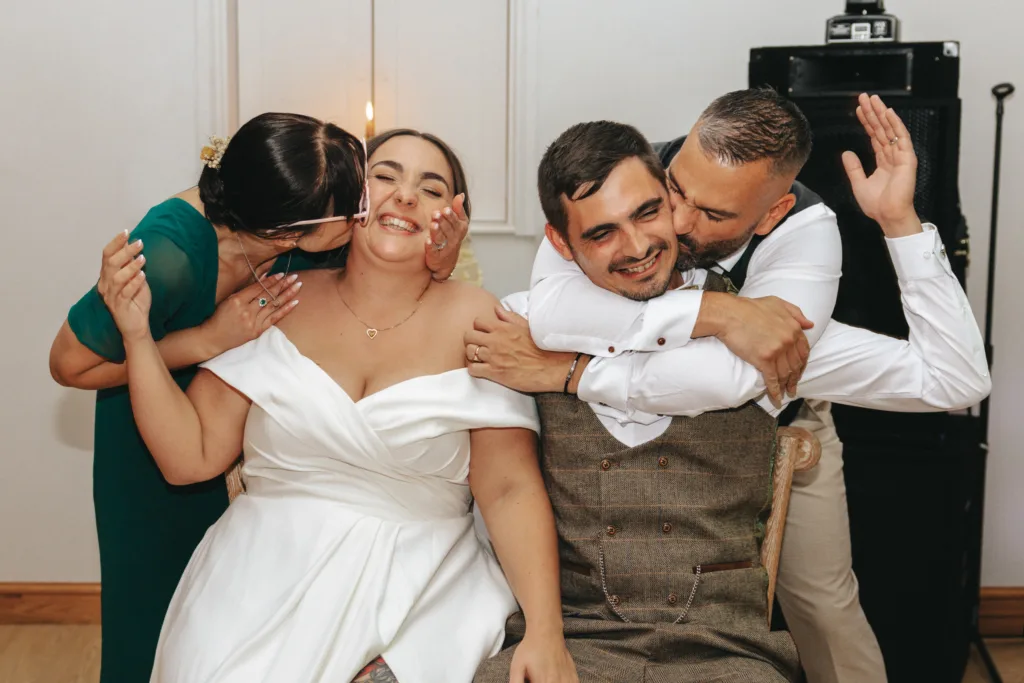 Two people in wedding attire sit smiling; the bride in a white dress and groom in a brown suit. In this joyful photo, a woman in green kisses the bride’s cheek, while a man hugs and kisses the groom. The atmosphere feels like a fun wedding photo game indoors. © Aimee Lince Photography - Wedding photographer in Lincolnshire, Yorkshire & Nottinghamshire. Photos from Photo Roulette, the photo dash game.