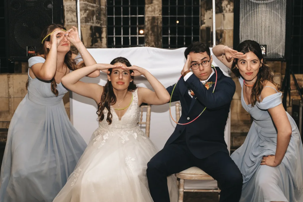 Four people pose for a fun photo indoors: a bride in white, groom in navy, and two bridesmaids in light blue dresses. All are making playful hand gestures, wearing glow stick necklaces as if mid-game, smiling in front of large speakers and a white backdrop. © Aimee Lince Photography - Wedding photographer in Lincolnshire, Yorkshire & Nottinghamshire