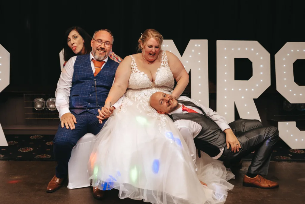 Four adults pose playfully for a photo in front of large illuminated "MR & MRS" letters. The bride in a white dress and groom rest on a sofa, laughing as their guests join in the fun as if it’s all part of a joyful game. © Aimee Lince Photography - Wedding photographer in Lincolnshire, Yorkshire & Nottinghamshire