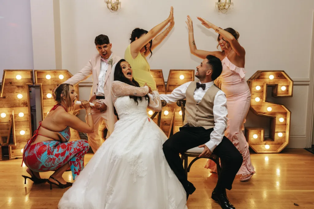 A bride and groom laugh joyfully as four guests, in colourful formal outfits, pose playfully around them for a photo in a brightly lit room. Large illuminated wooden letters spelling “LOVE” add a festive, game-like atmosphere to the wedding celebration. © Aimee Lince Photography - Wedding photographer in Lincolnshire, Yorkshire & Nottinghamshire