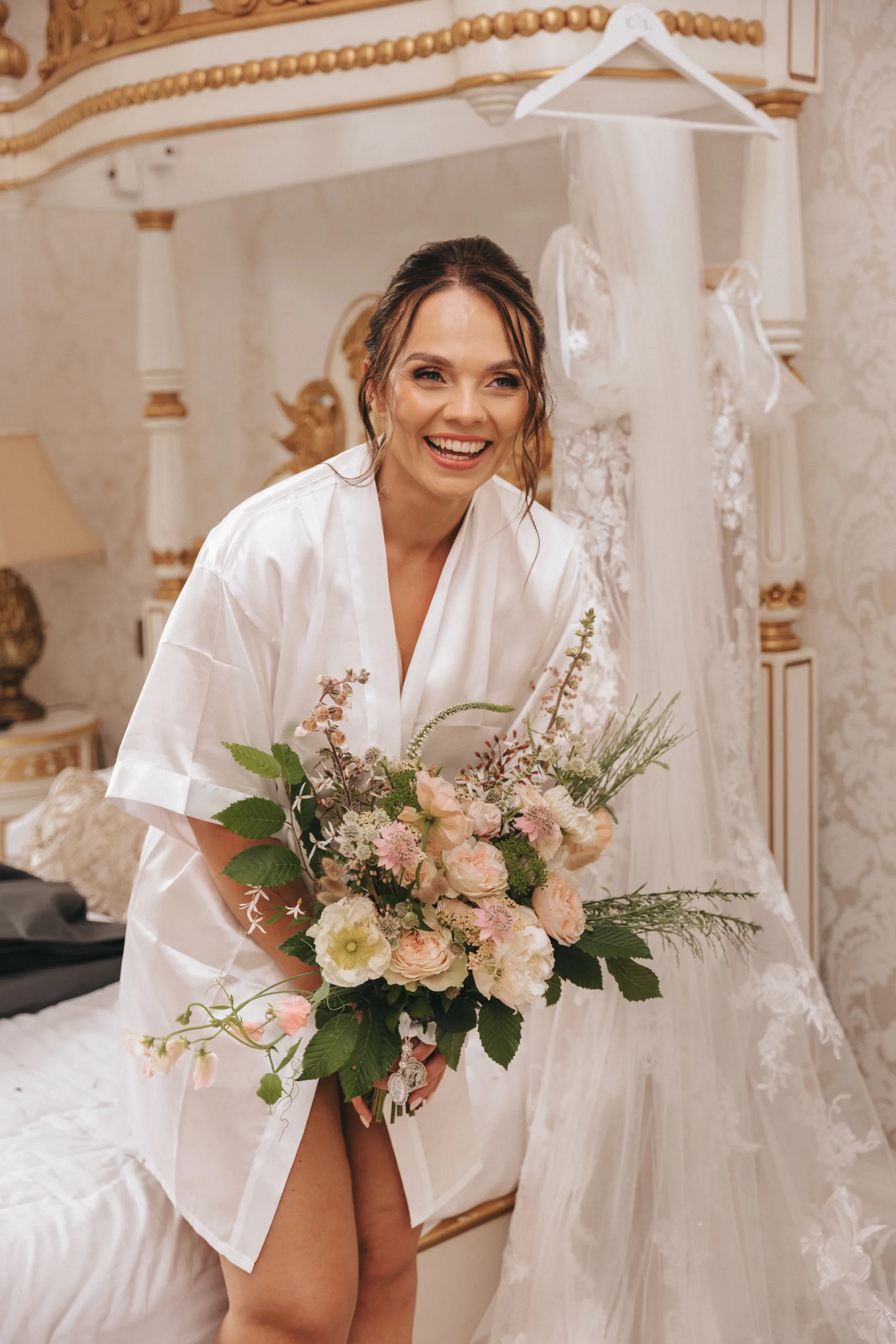A bride in a white robe holding a bouquet in front of her bed captured by a photographer.