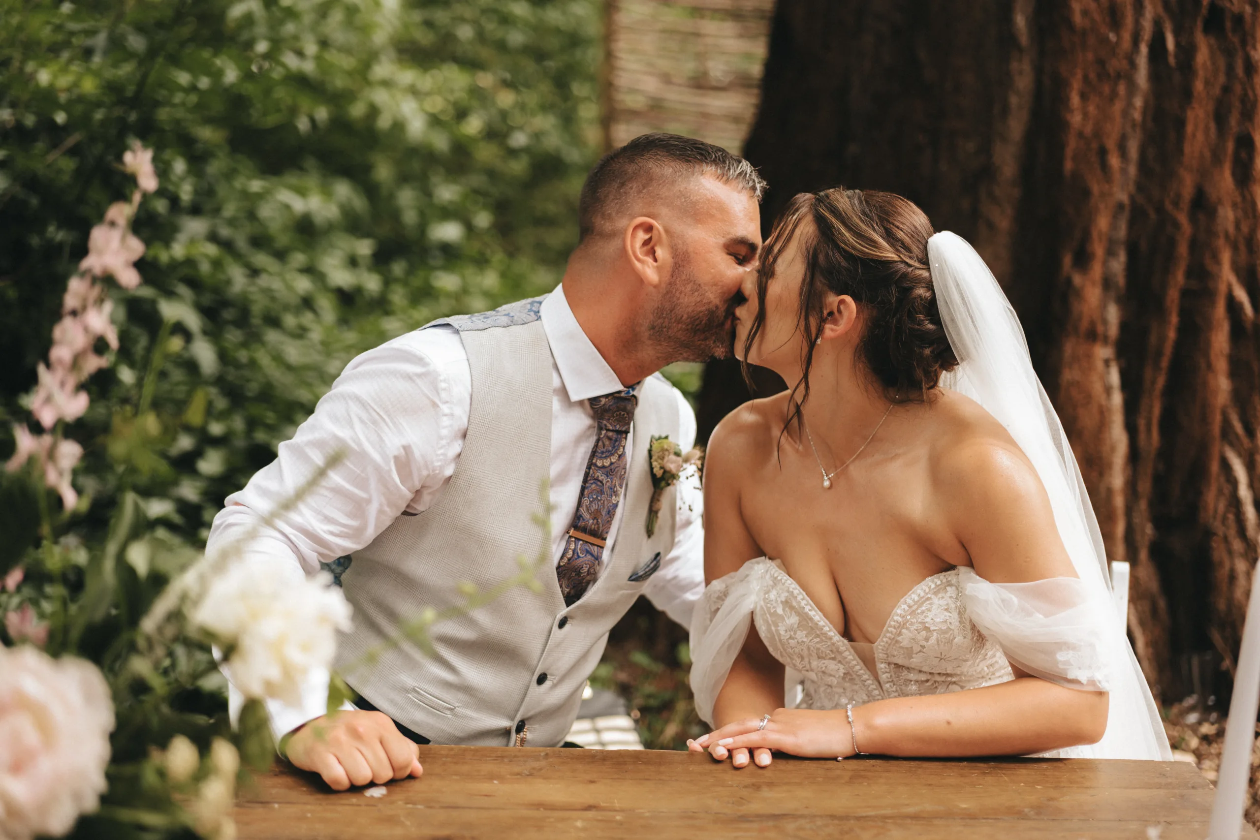 A bride and groom kissing at a wooden table in a picturesque setting, captured by a Lincolnshire photographer.