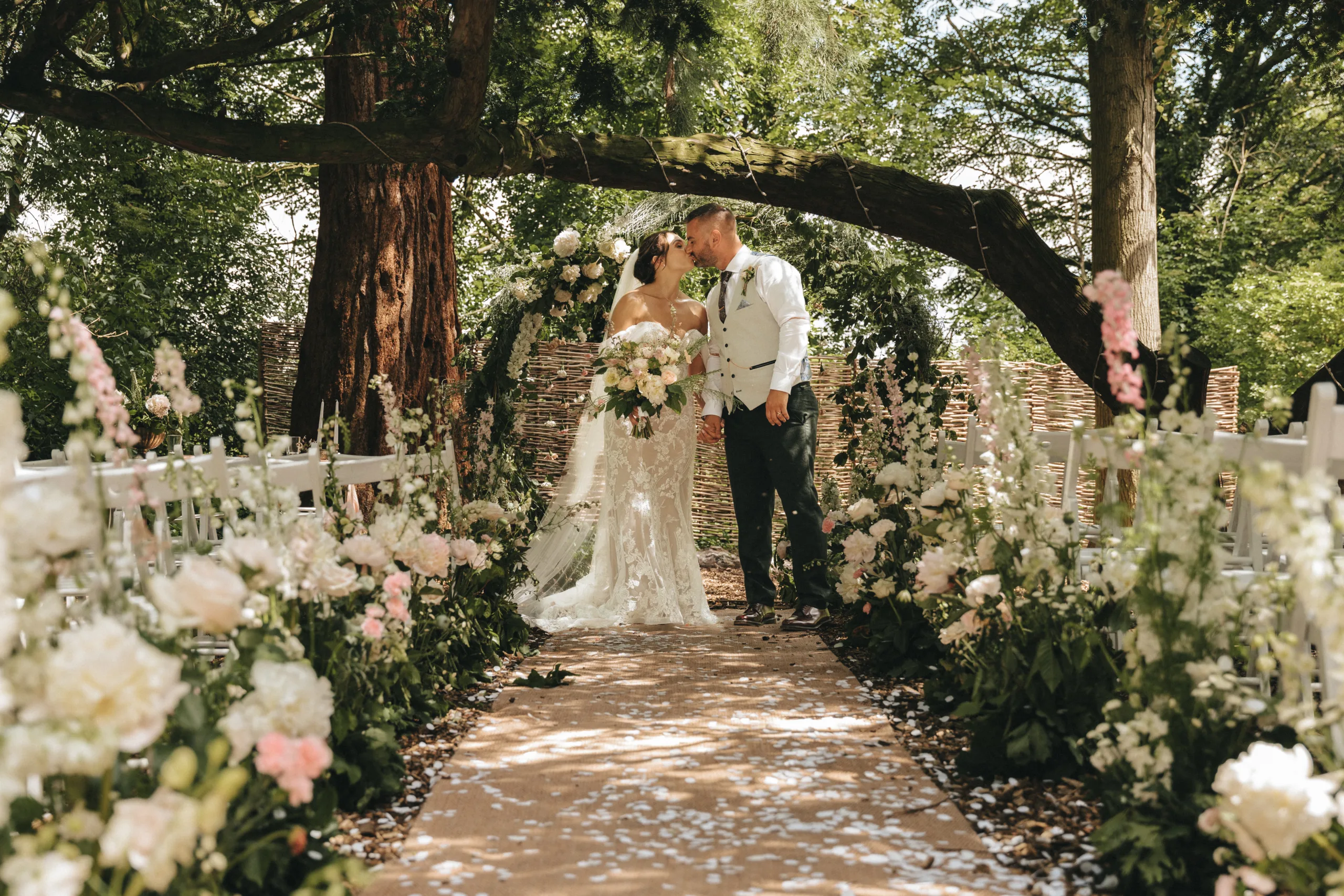 A bride and groom kissing under a tree in a garden, captured by a photographer.