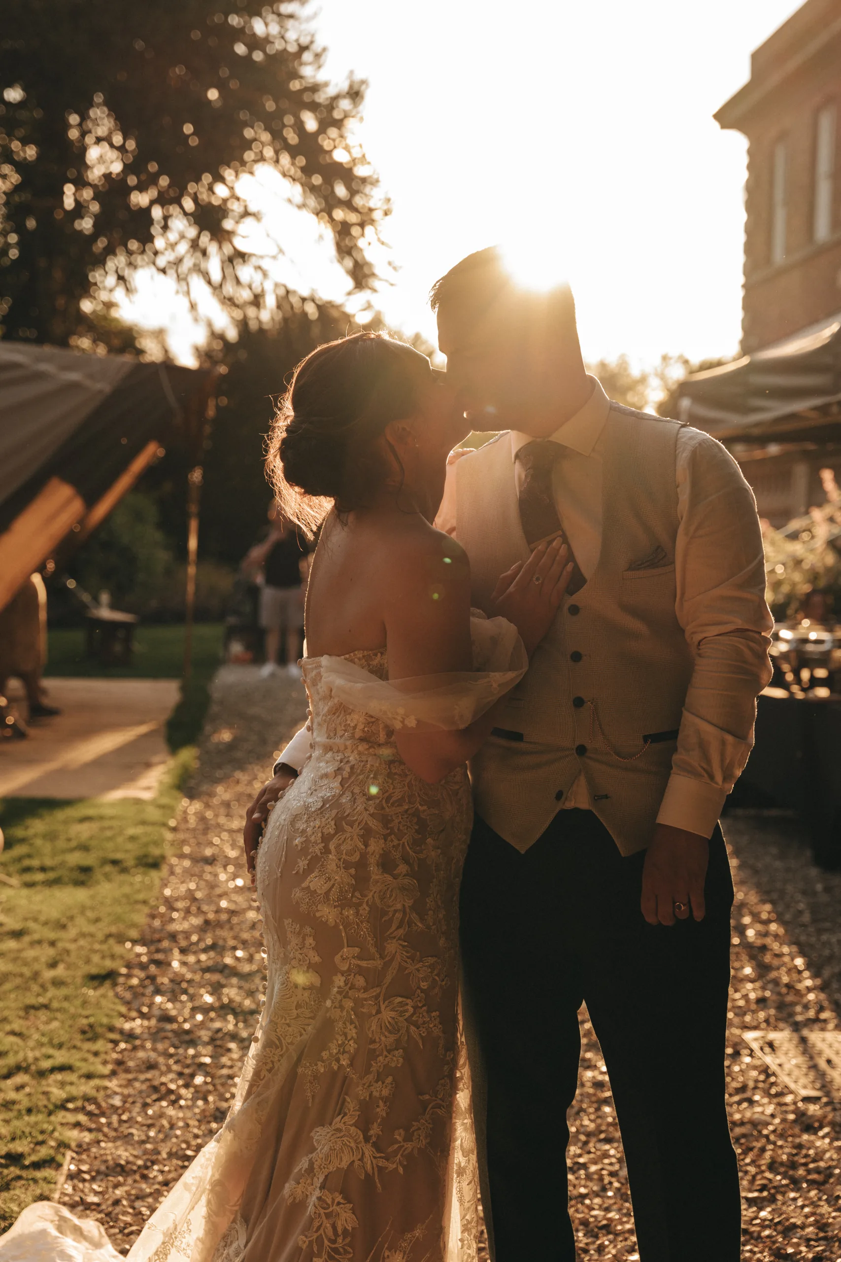 A bride and groom kiss in front of a tent at sunset while a photographer captures the moment.