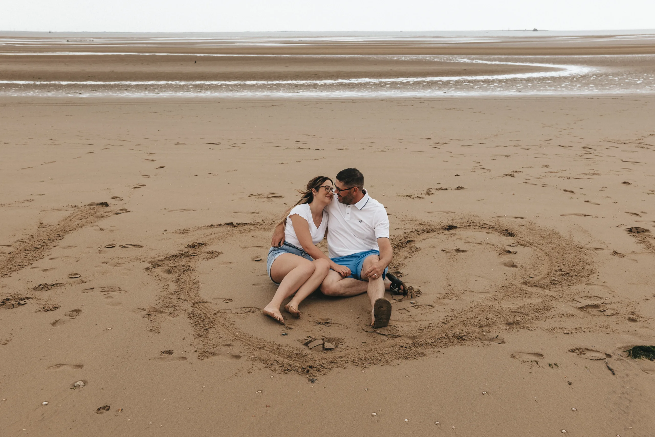 An couple sits on the sand during an engagement shoot on Cleethorpes beach in a heart shape. A photographer captures the moment.