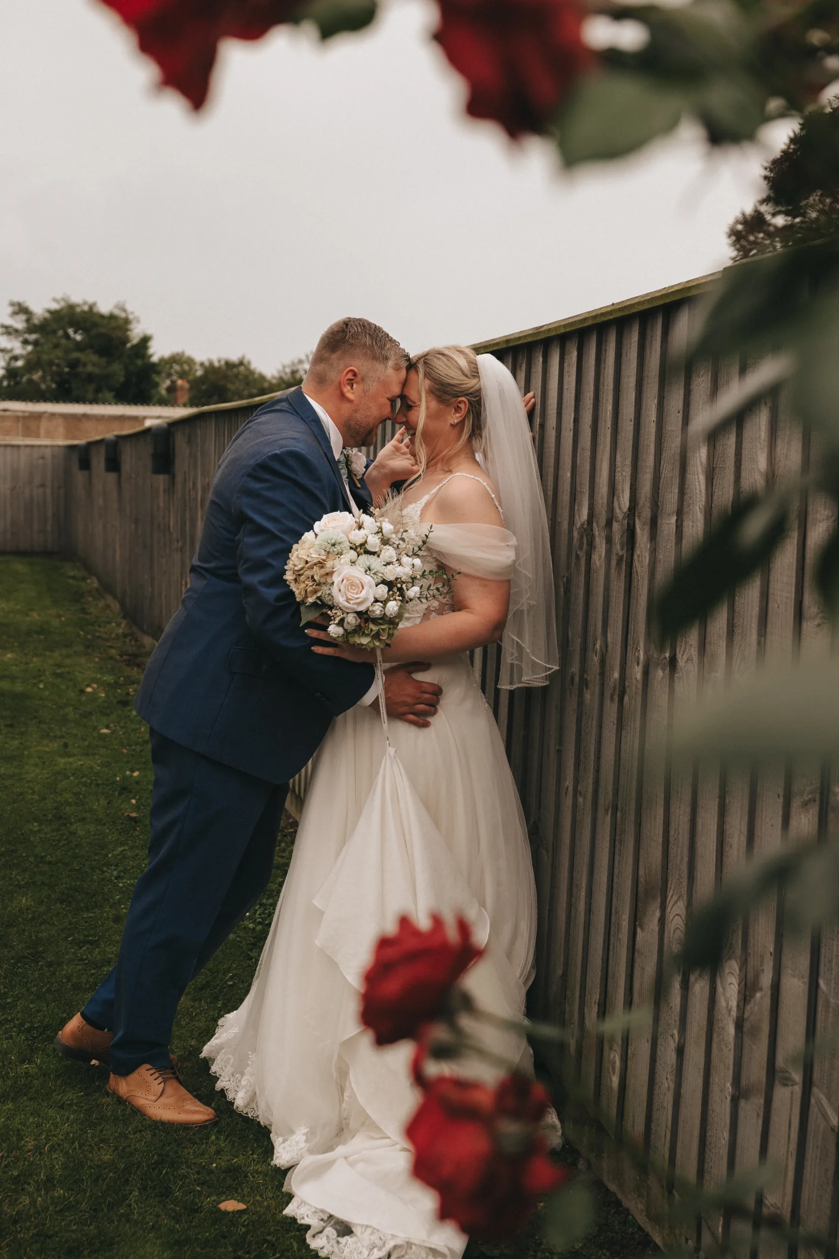 A bride and groom kissing in front of a fence with red roses, captured by a photographer.