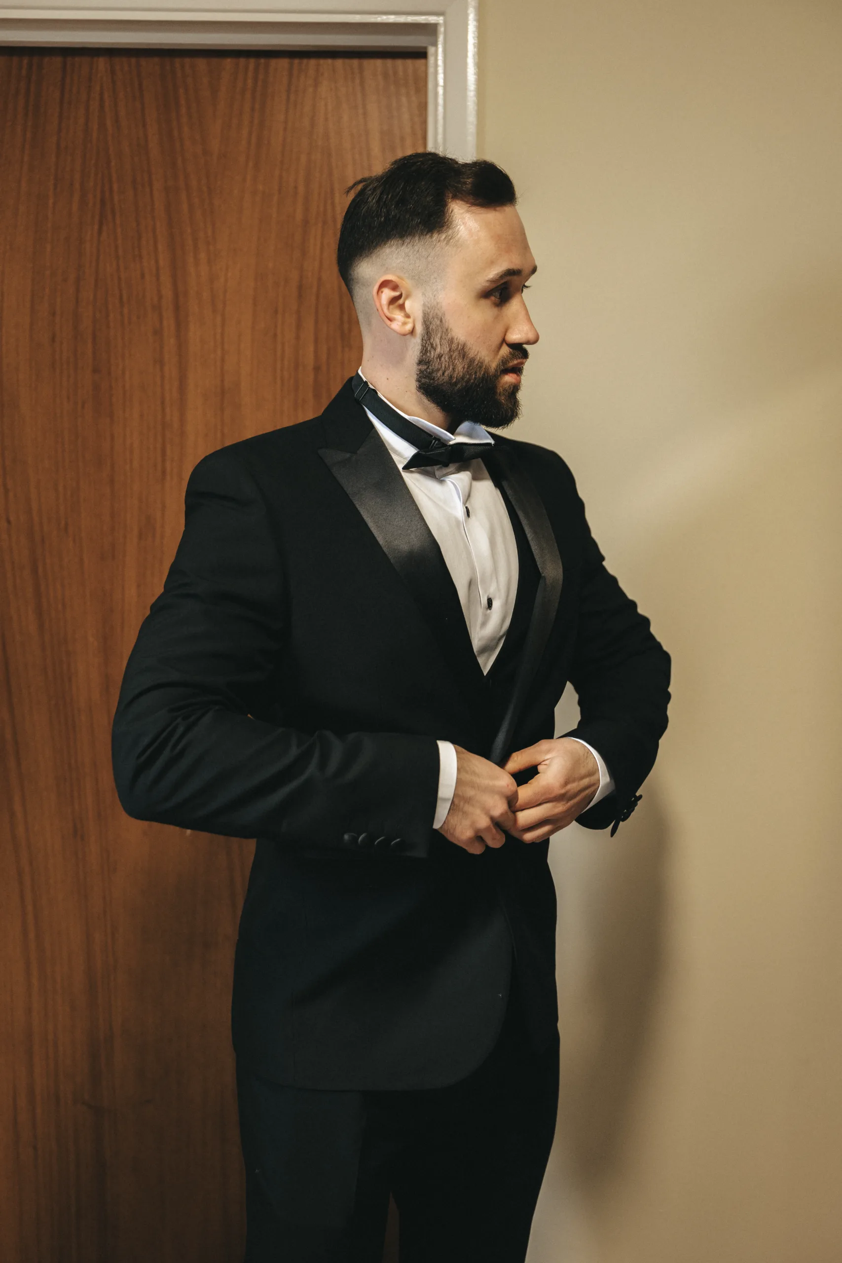 A man in a tuxedo is standing in front of a door, posing for the photographer.