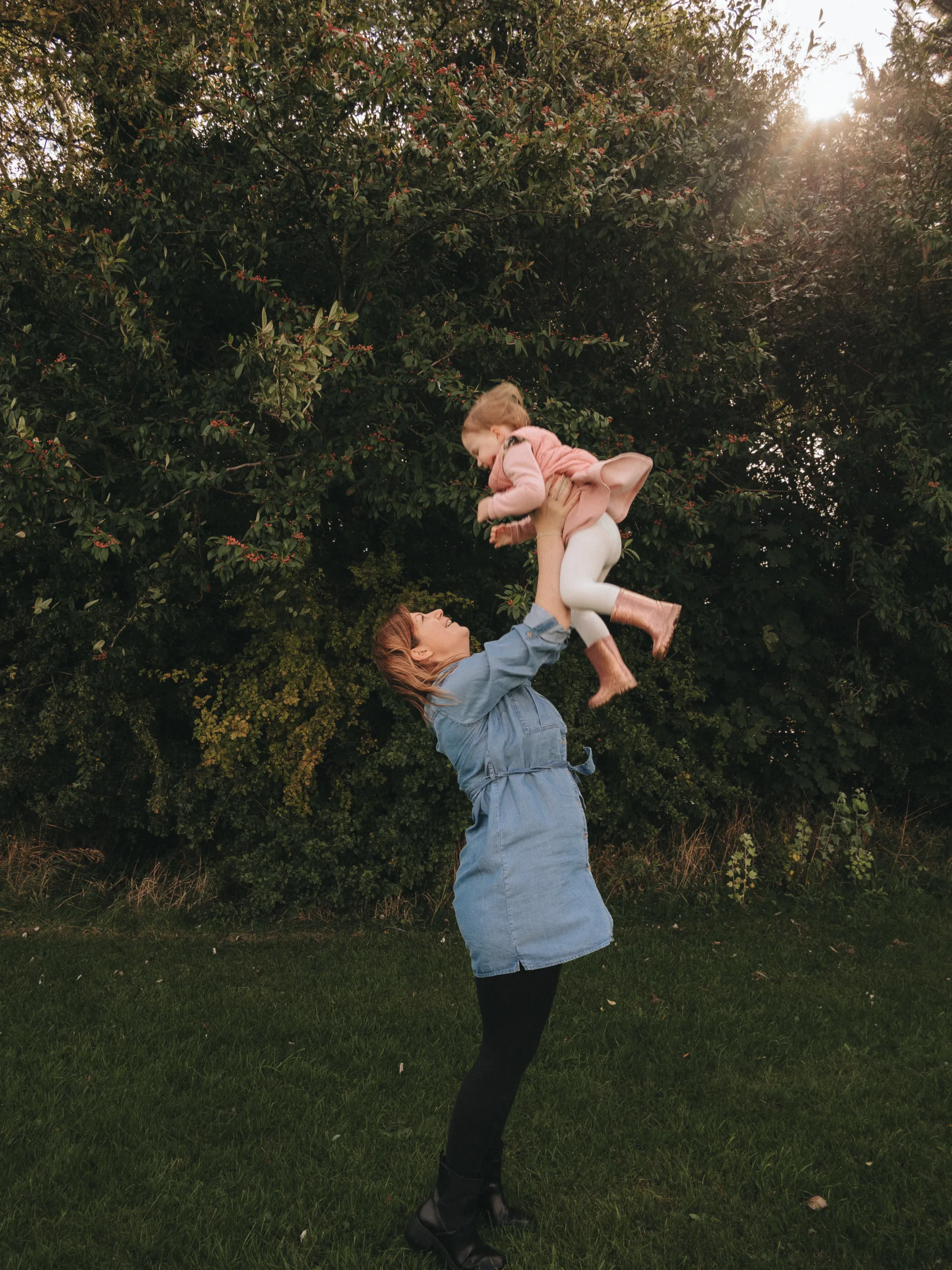 A woman is holding her daughter in the air in a field, captured by a photographer.