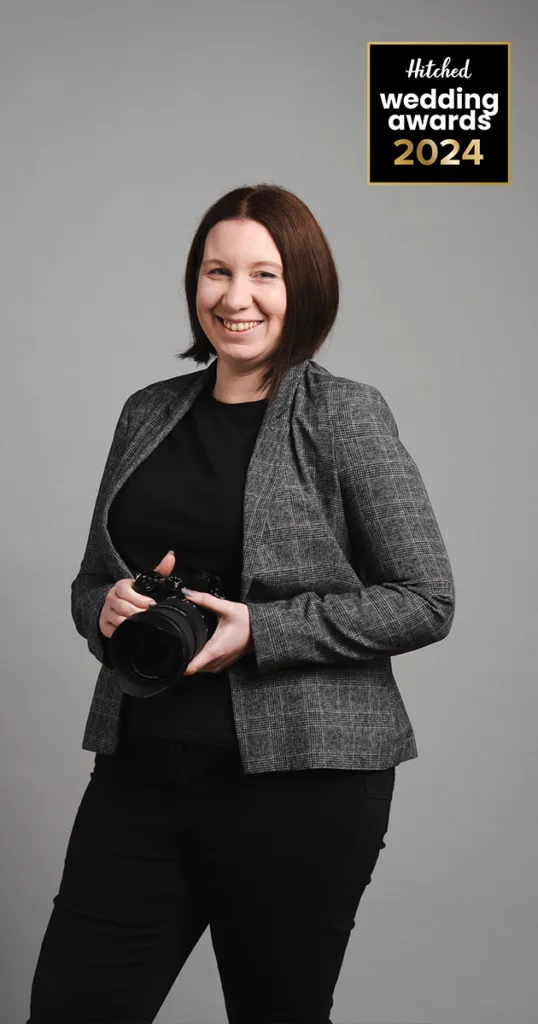 A person with shoulder-length brown hair smiles while holding a camera, embodying an award-winning wedding photographer. They wear a gray plaid blazer over a black shirt and trousers, standing against a light gray background. In the top right corner, a logo reads "Hitched Wedding Awards 2024" in black, gold, and white. © Aimee Lince Photography - Wedding photographer in Lincolnshire, Yorkshire & Nottinghamshire