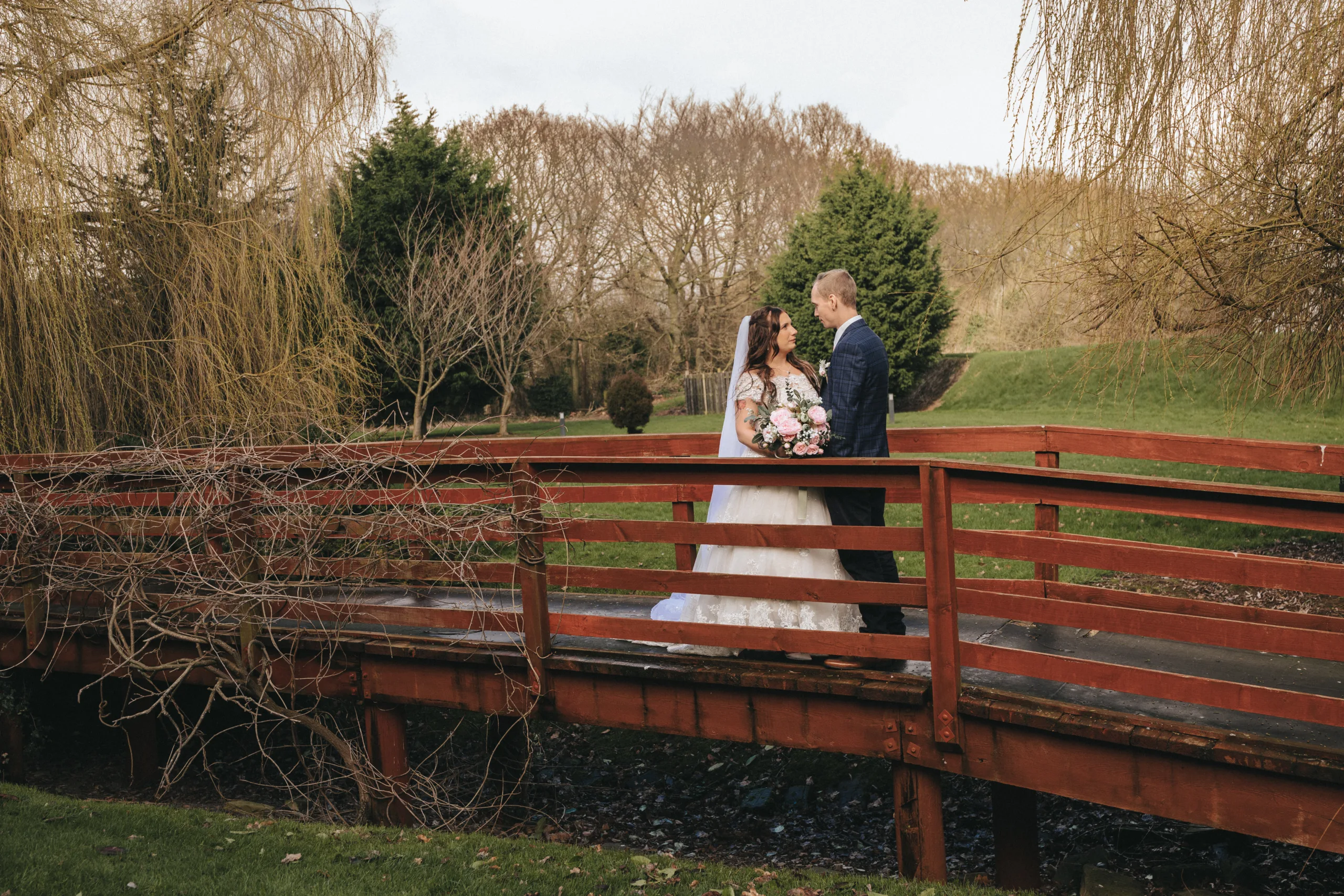 A bride and groom standing on a bridge in a field.