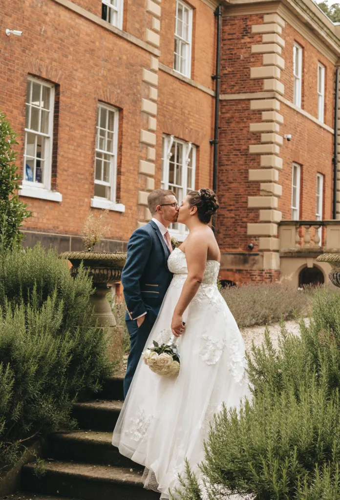 A newlywed couple shares a kiss on stone steps beside manicured shrubbery, with a classic brick building in the background, symbolizing a romantic celebration of their union.