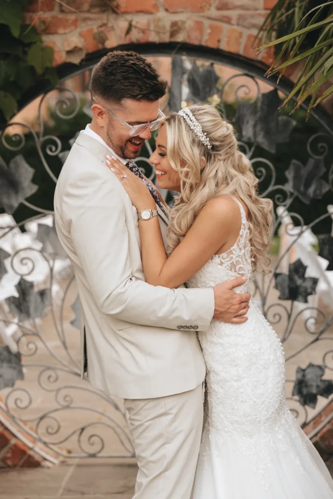 A joyful bride and groom share a loving embrace and laughter against a decorative backdrop, their wedding attire radiating elegance and bliss on their special day.