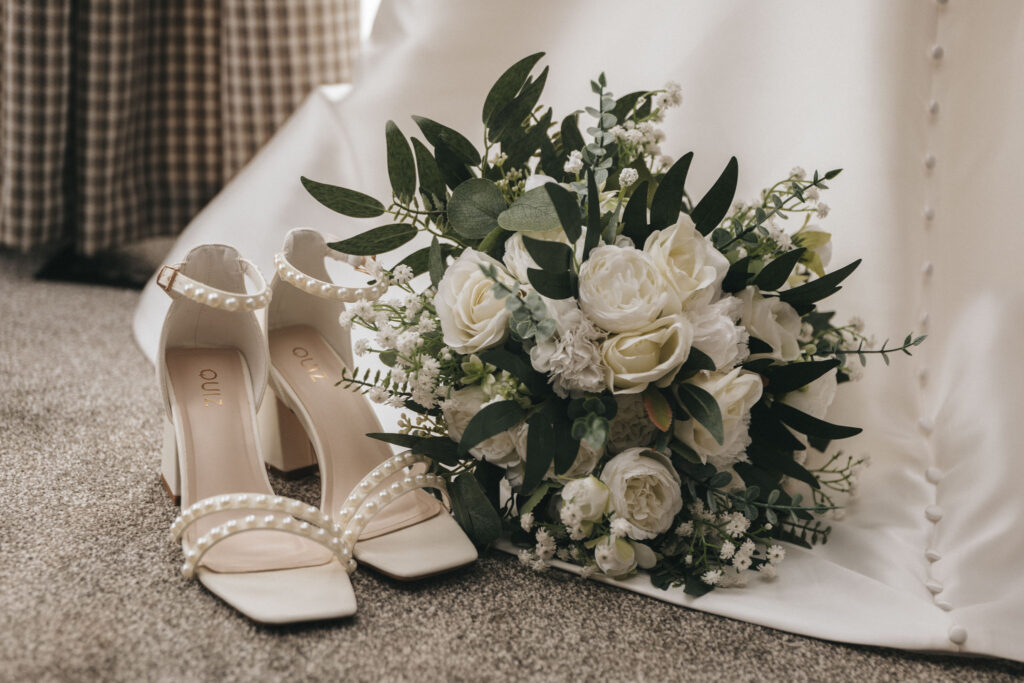 A pair of elegant white high-heeled shoes with pearl embellishments rests on a carpeted floor next to a bouquet of white roses and greenery. Captured by a talented wedding photographer, the scene unfolds in front of a checkered fabric and white curtain, evoking a soft, bridal atmosphere. © Aimee Lince Photography - Wedding photographer in Lincolnshire, Yorkshire & Nottinghamshire