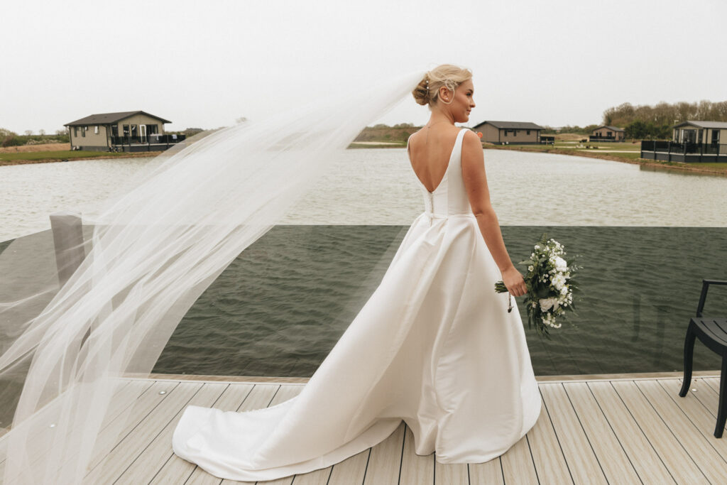 A bride stands on a wooden deck overlooking a lake at the Brackenborough Hotel, wearing a long white dress with a low back and a flowing veil. She holds a bouquet of white and green flowers, her hair styled in an elegant updo. In the background, quaint houses and trees dot the scenery. © Aimee Lince Photography