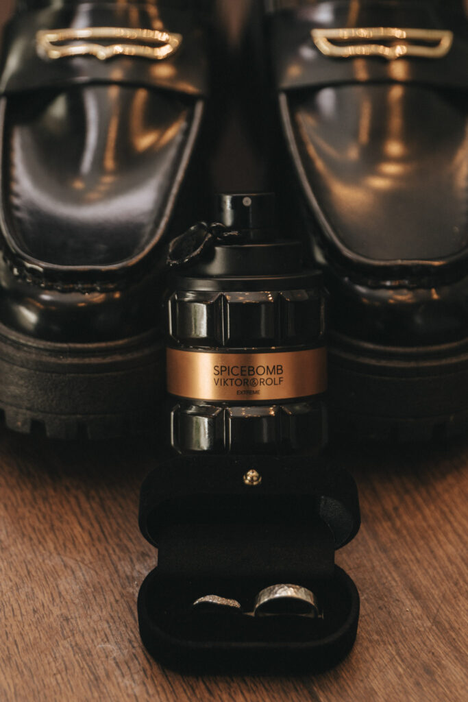 A black bottle of Spicebomb by Viktor & Rolf is placed on a wooden surface, flanked by glossy black leather loafers. In front, an open black velvet ring box cradles a silver ring. This elegant scene perfectly captures the essence of a sophisticated wedding day. © Aimee Lince Photography