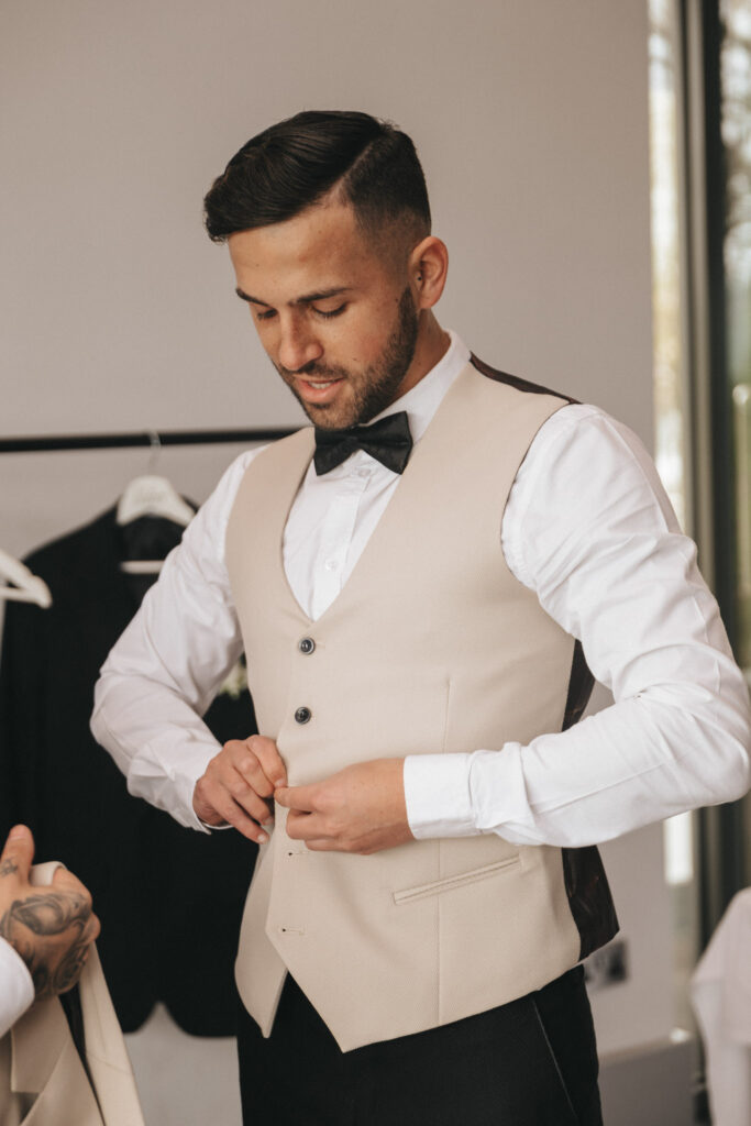 A man with short dark hair and a beard is buttoning his beige vest over a white shirt, preparing for a wedding. He wears a black bow tie and stands next to a clothing rack with a black jacket on it. A hand with tattooed fingers holds the vest. Sunlight pours in through the room's large window. © Aimee Lince Photography - Wedding photographer in Lincolnshire, Yorkshire & Nottinghamshire