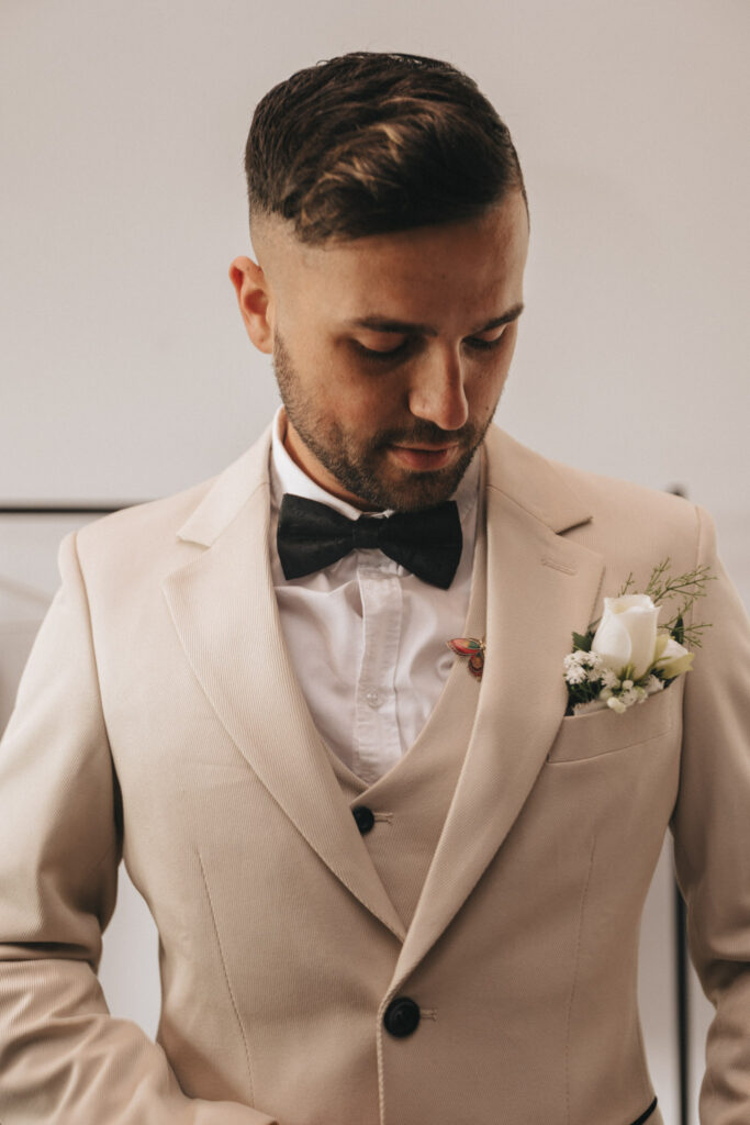 A man in a beige suit with a white shirt and black bow tie looks down, embodying the essence of marriage. He has short hair, a trimmed beard, and wears a white flower boutonniere on his jacket. The background is simple and out of focus, enhancing his presence in the photo captured at Brackenborough Hotel. © Aimee Lince Photography