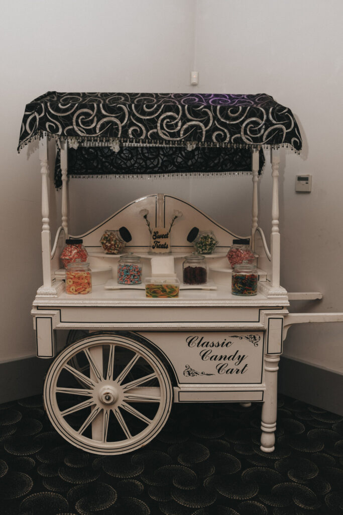 A vintage candy cart with a black and white ornate canopy graces the elegant setting of a Brackenborough Hotel wedding. Filled with jars of colorful candies, the cart gleams with decorative writing reading "Classic Candy Cart" and "Sweet Treats," perfect for delighting the bride and her guests. © Aimee Lince Photography
